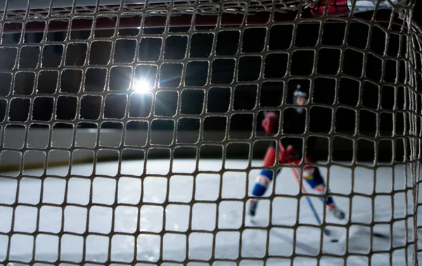 View from behind the net of the gate at a male hockey player scoring a goal. An athlete in uniform and skates plays hockey on a dark ice arena. Hockey player hits the puck with a stick. Close up. — Zdjęcie stockowe