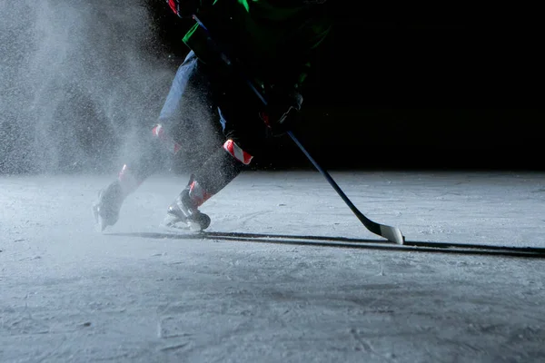 Hockeyspelare träffar pucken med pinne på svart bakgrund av isarenan. En manlig idrottsman i uniform och skridskor spelar hockey på mörka stadion rinken. Träffar pucken, en explosion av pulversnö. Närbild. — Stockfoto
