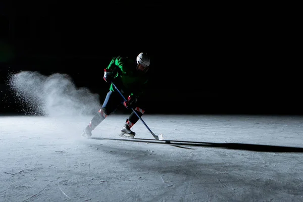 Hockey player hits the puck with stick on a black background of the ice arena. A male athlete in uniform and skates plays hockey on dark stadium rink. Hitting the puck, an explosion of powdered snow. — Zdjęcie stockowe