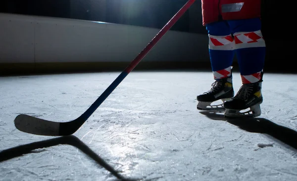 Man hockeyspelare i sportuniform och skridskor står på isarena med pinne i händerna. En idrottsman hones sina färdigheter i att spela hockey på isen av stadion i mörker med bakgrundsbelysning. Ben på nära håll. — Stockfoto