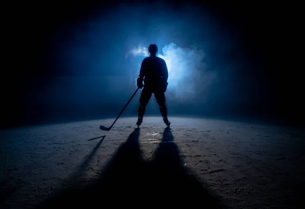 Dunkle Silhouette eines männlichen Eishockeyspielers in Uniform, Helm und Schlittschuhen mit Stock auf der Eisarena mit Rauch und blauem Gegenlicht. Sportler posiert auf der Eisbahn des Stadions. Wintersport. — Stockfoto