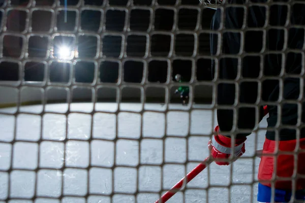 View from behind the net of goalkeeper of the hockey player preparing to hit the goal. An athlete in uniform and skates plays hockey on dark ice arena. Hockey player hits puck with stick. Close up. — Zdjęcie stockowe