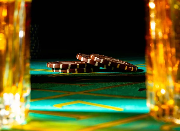 Brown casino chips lie on a gaming green table between two glasses of whiskey. Chips on a roulette table close up on a black background. Alcohol, gambling, entertainment, money.