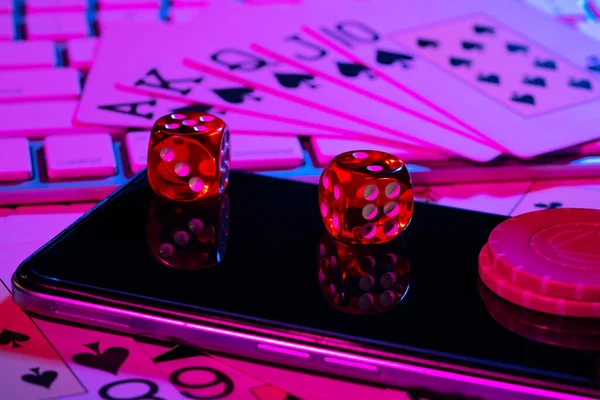 Cards, phone, casino chips and dice close up. Computer keyboard and smartphone, illuminated with pink light. Concept of gambling, online betting in casino. Play poker over internet. Online gambling. — Foto de Stock
