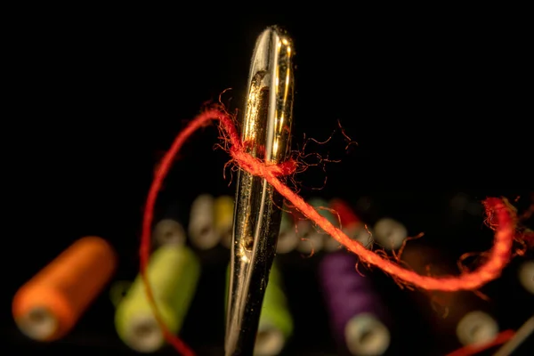 Œil d'aiguille fileté avec un morceau de fil rouge sur fond noir avec des bobines de fil colorées. Aiguille tailleur en acier avec filetage de perméation. Outil de couture, broderie, réparation de vêtements. — Photo