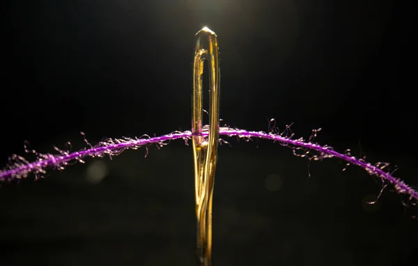 Macro image of a needle eye threaded with piece of purple thread on black background. Steel shiny tailors needle with permeation thread close up. The tool for sewing, embroidery, repair of clothes. — Foto Stock