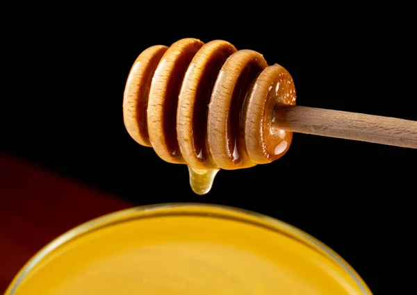Honey dripping, pouring from honey dipper into glass bowl on black background. Healthy organic thick honey dipping from wooden honey spoon, close up. Golden liquid, sweet molasses, sugar syrup.