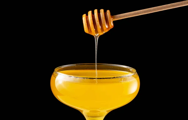 Honey dripping, pouring from honey dipper into glass bowl on black background. Healthy organic thick honey dipping from wooden honey spoon, close up. Golden liquid, sweet molasses, sugar syrup.