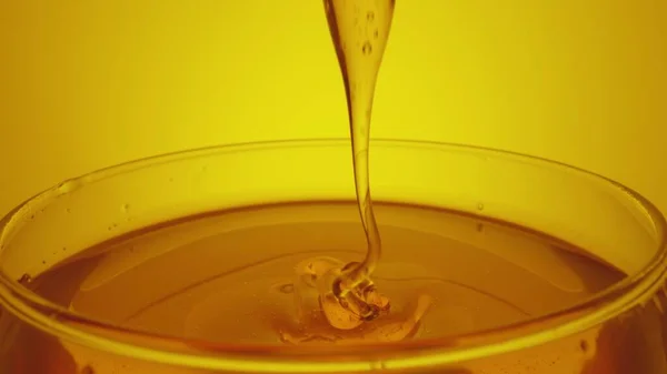 Honey dripping, pouring from spoon in glass. Thick honey molasses dripping into full glass. Close up of golden honey liquid, sweet product of beekeeping. Sugar syrup is pouring on yellow background. — Stockfoto