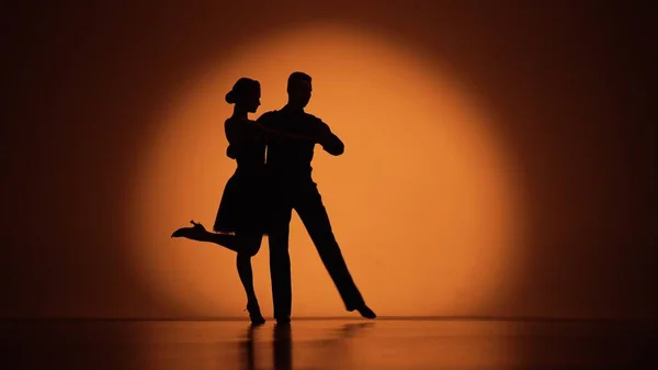 Couple of dancers approach each other and begin to dance a passionate Argentine tango. Elements of latin ballroom dance choreography in studio with orange brown background. Dark silhouettes of a man