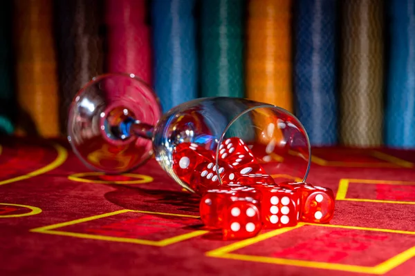 Fallen glass of champagne with red dice scattered on table against backdrop of casino chips. Close up of set of dice for craps, roulette, poker on a red gaming table in a casino. Gambling background