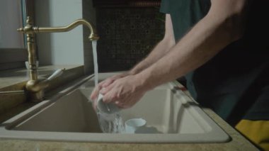 Erkek elleri mutfakta beyaz bardakları yıkıyor. Eski bir musluktan lavaboya dökülen bir su deresini kapatın. Bulaşık, bulaşık yıkama. Ev işi, temizlik, temizlik.