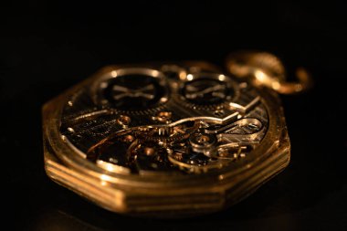 Siyah masa yüzeyinde duran, saat mekanizması açık olan eski altın cep saatinin arka görüntüsü. Altın antika saat mekanizması, dişli tekerlekler, dişliler, dişliler ve vidalar. İçerideki sekizgen retro saat