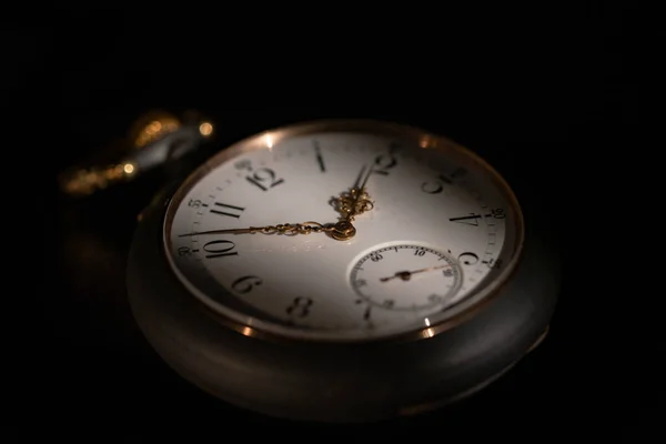 Round white dial of an antique clock with golden second, minute and hour hands on a black blurred background. Black retro mechanical pocket watch in the dark. Luxury aged timepiece. The play of light