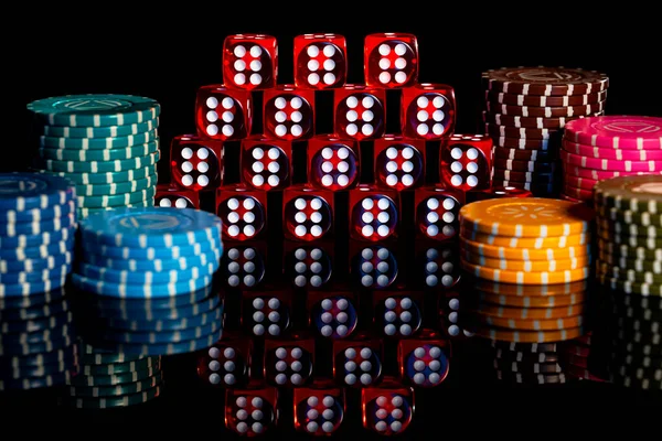 Pile of red dice and stack of casino chips on isolated black background. Pyramid of dice with number six and colored chips on a reflective table surface. Background of gambling in the casino. Craps