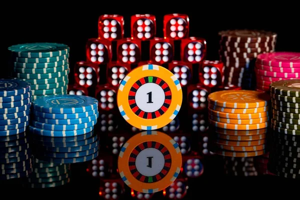 Pile of red dice and stack of casino chips on isolated black background. Pyramid of dice with sixes and chip with unit on reflective table surface. Background of gambling in casino. Craps, poker