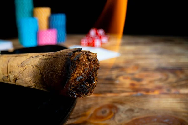 Smoking Cuban cigar against the backdrop of casino chips, cards and dice lying on wooden table. Burning Cuban cigar on an isolated black background in casino. Concept of gambling, luxury, success