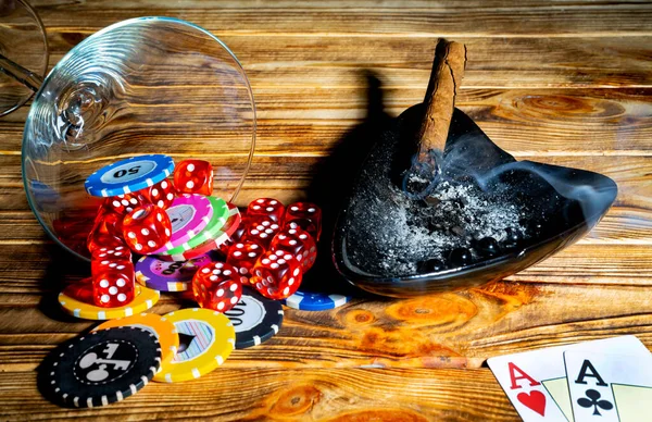 Burning cigar, cards, set of dice and casino chips in a fallen glass on a wooden table. Two aces, chips different denominated and red dice. A smoking Cuban cigar in a black ashtray. Gambling