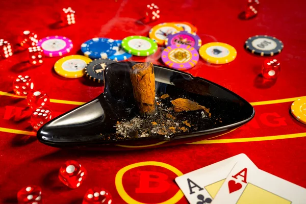 Smoking cigar in an ashtray, cards, chips and dice on a red gambling table in a casino. Two aces, chips different denominated for playing poker, bridge, blackjack. Gambling background. The concept of