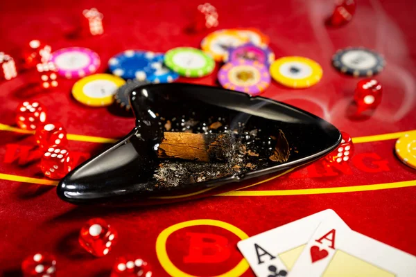 Smoking cigar in an ashtray, cards, chips and dice on a red gambling table in a casino. Two aces, chips different denominated for playing poker, bridge, blackjack. Gambling background. The concept of