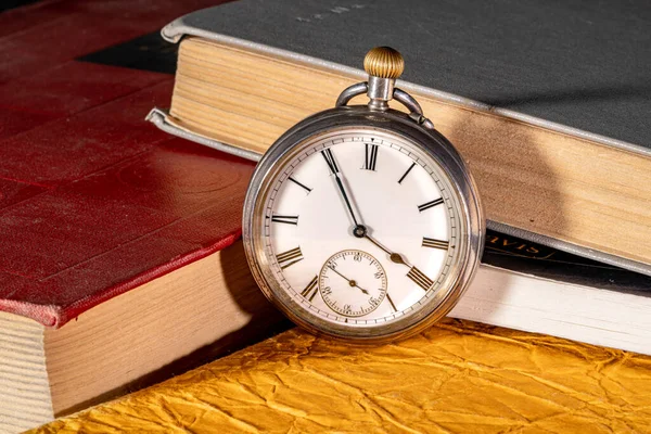 Silver vintage clock on the background of old books. Round retro clock near red, gray and gold book covers in the library. The concept of time, past, knowledge, memories