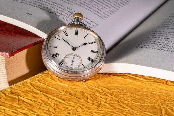 Antique silver pocket watch on background of an open book with text. Round vintage clock near old books with red and yellow covers in library. The concept of time, learning, knowledge, past, memories