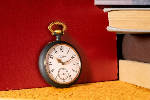 Antique silver retro clock on background of stacks of books in library. Round old vintage clock with dial and golden hands near books with red, yellow and gray covers. Concept of time, education