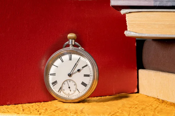 Antique silver retro clock on background of stacks of books in library. Round old vintage clock with dial and hands near books with red, yellow and gray covers. Concept of time, education, knowledge