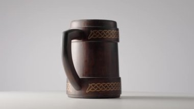 Açık gri stüdyo arka planında dönen saplı tahta meşe kupası. Süslü kahverengi kupa. Bira ya da kvass için el yapımı ahşap tankard. Kırsal bir geçmişi var. Barda ya da barda bir şeyler içmek için.