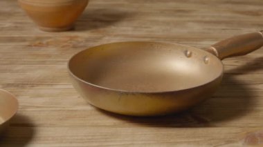 An old copper frying pan with a handle on a rustic wooden table next to an clay jug. Gold round empty skillet, stainless or aluminum pan with handle. Kitchenware, utensils for cooking and frying