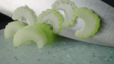 Male hands cutting celery stalks with knife into small slices. Pieces of juicy raw chopped celery on gray plastic board on a restaurant kitchen table. Chef cooking vegan salad with raw healthy