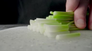 Green onion are cut with knife in kitchen. Mans hand with sharp knife cutting the stalks of scallion or shallot on gray plastic board. Chef restaurant cooking vegan salad with raw healthy vegetables