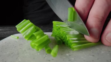 Green onion are cut with knife in kitchen. Mans hand with sharp knife cutting the stalks of scallion or shallot on gray plastic board. Chef restaurant cooking vegan salad with raw healthy vegetables