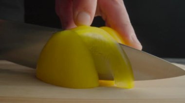 Hands of man with knife slicing yellow sweet pepper into semiring. Process of cutting bell peppers in slices on wooden board on dark background. Preparation of salad from raw vegetables. Healthy food