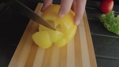 Male hands with a knife cutting yellow sweet pepper into pieces. Slicing bell pepper on a wooden board. On a dark table lie a red tomato and green lettuce for making fresh salad. Raw organic healthy