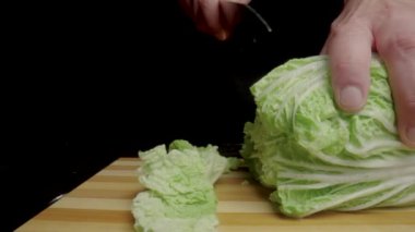 Male hands cutting Chinese cabbage or green lettuce leaves with sharp knife. Chef slicing leafy greenery on wooden cutting board. Cooking in the kitchen or in a restaurant. Black background