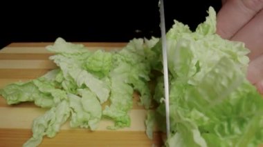Male hands cutting Chinese cabbage or green lettuce leaves with sharp knife. Chef slicing leafy greenery on wooden cutting board. Cooking in the kitchen or in a restaurant. Black background