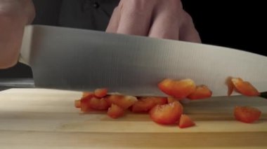 Hands of man with knife dicing red sweet pepper. Process of cutting bell peppers in pieces or slices on wooden board. Preparation of salad from raw vegetables in kitchen or restaurant on dark