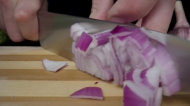 A mans hand cutting half an onion with a knife. Chopping an bulb on a wooden board, dicing a red onion. Onion, green lettuce and garlic on a black studio background. Cooking a healthy fresh salad of