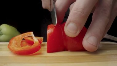 Hands of man with knife cutting red sweet pepper on black background. Process of slicing bell peppers in pieces or slices on wooden board. Red, yellow sweet peppers and garlic. Preparation of salad