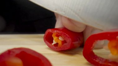 Male hands slicing red hot chili pepper with sharp knife on black background. Chef cutting spicy peppers with seeds into thin slices on wooden board. Cooking in kitchen or in restaurant. Healthy