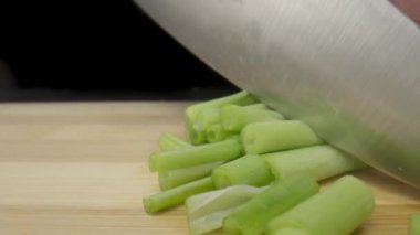 Green onion are cut with knife in kitchen on black background. Mans hand with sharp knife cutting the stalks of scallion or shallot on wooden board. Chef restaurant cooking vegan salad with raw