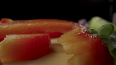 Red sweet pepper slices and green onion stalks on wooden board. Pieces of ripe raw peppers, shallot and garlic on black background. Ingredients for fresh vegetable salad. Macro shot. Slow motion ready