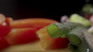 Red sweet pepper slices and green onion stalks on wooden board. Pieces of ripe raw peppers, shallot and garlic on black background. Ingredients for fresh vegetable salad. Macro shot. Slow motion ready