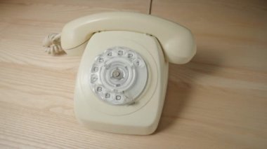 Old white rotary telephone with twisted cord on beige wooden table. Retro landline phone with rotary dialer and handset. Vintage analog plastic rotary telephone with dialing disk. Concept of