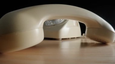 Old white rotary telephone with removed handset lying on a beige wooden table. Retro landline phone with dialing disk and twisted cord on black background. Vintage analog plastic phone. Concept of