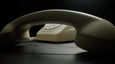 Old white rotary telephone with removed handset lying on dark wooden table. Retro landline phone with dialing disk and twisted cord on black background. Vintage analog plastic phone. Concept of