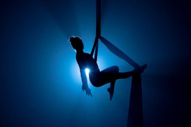 A young woman performing in a circus on aerial silk in the dark with blue light. A female equilibrist balancing on a height. Performance of aerial gymnast. For sports, acrobatic, circus school.
