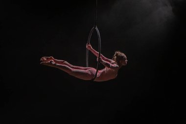 Aerial acrobat in the air ring. Young woman performs the acrobatic elements in the air hoop. Aerialist in on black background dark studio with backlight and smoke. For sports, acrobatic, circus school