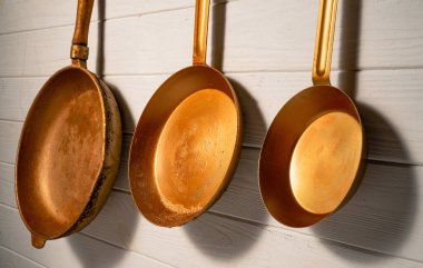 Three rustic copper fryingpans hanging on white kitchen wall in restaurant or at home. Round empty gold or yellow pans with handles. Kitchenware, utensils for cooking and frying, interior decoration